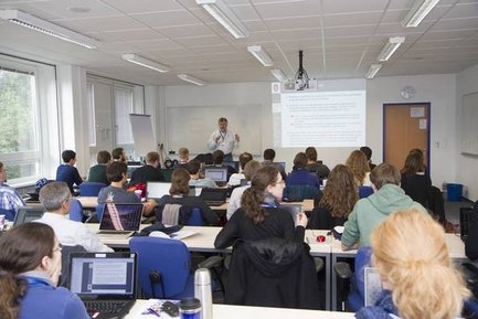 Seminarraum mit mehreren Personen, die an Tischen sitzen und einem Vortragenden zuhören, der vor einer Projektionsfläche steht.