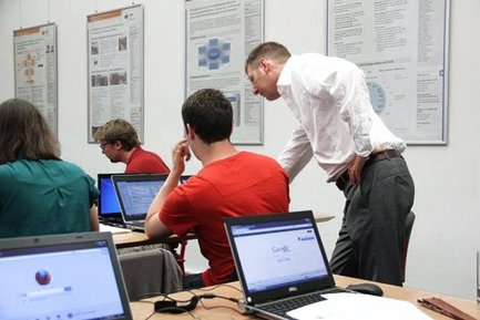 Mehrere Personen sitzen an Tischen mit geöffneten Laptops, eine Person steht und beugt sich vor, um etwas zu erklären. Im Hintergrund hängen Informationsplakate an der Wand.