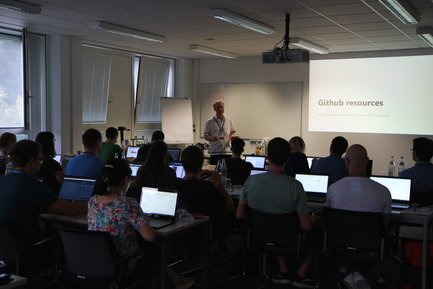 Ein Mann steht vor einem hell erleuchteten Projektionsbildschirm mit dem Text 'Github resources'. Er spricht zu einem Publikum, das an Tischen sitzt und auf Laptops arbeitet.
